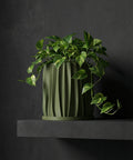 A green planter pot with a plant sits on a wooden table in a room with a rug and a basket hanging on the wall.