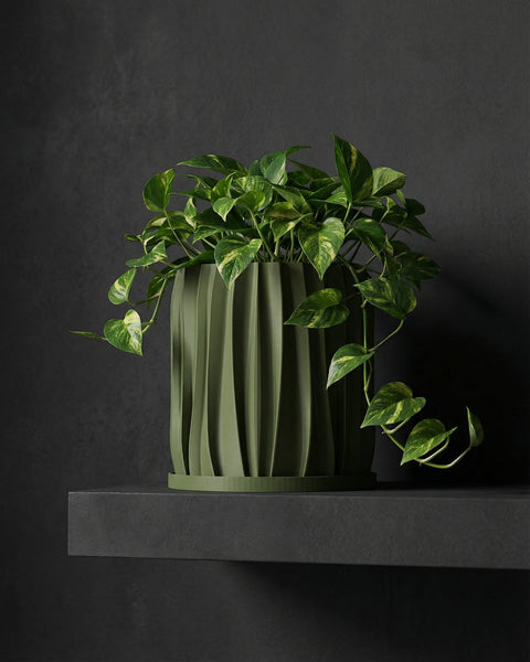 A green planter pot with a plant sits on a wooden table in a room with a rug and a basket hanging on the wall.