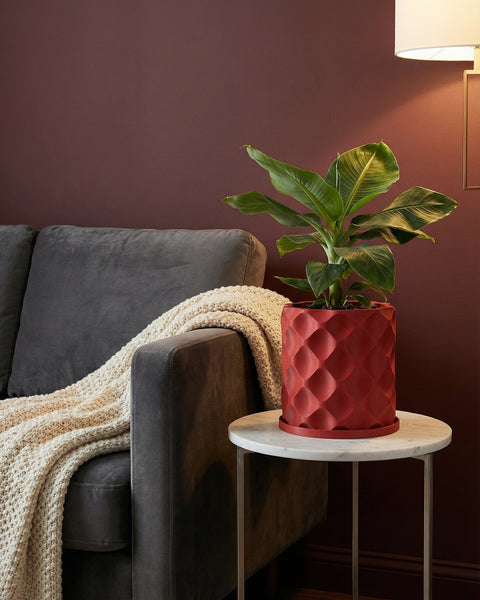 A red planter pot with a geometric design sits on a marble table next to a gray couch with a white blanket.