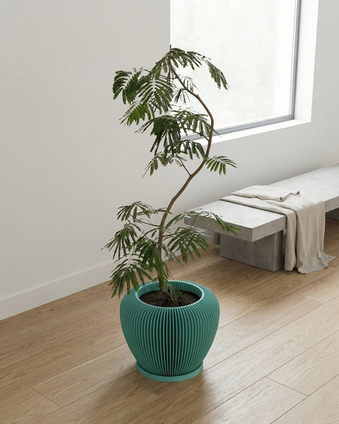 A teal-colored planter pot with a plant inside sits on a wooden floor next to a bench.