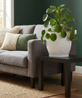 A Pilea peperomioides plant sits in a white, textured planter on a dark wooden side table next to a gray couch.