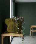 A green planter pot with a plant on a white table, next to a white chair and a black and white painting.
