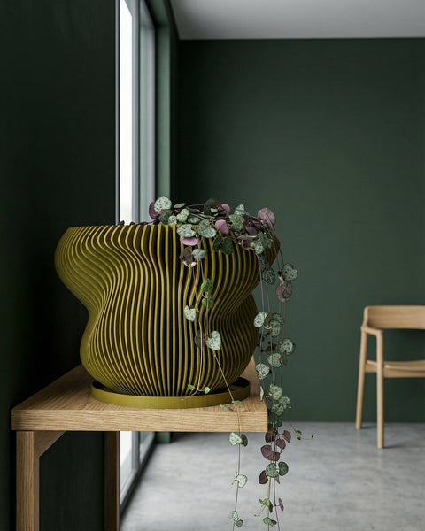 A green planter pot with a plant on a white table, next to a white chair and a black and white painting.