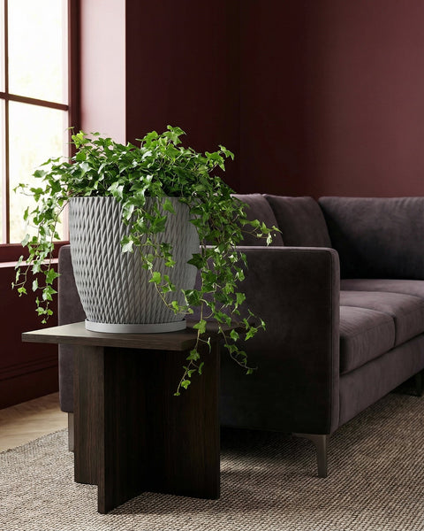A gray planter pot with a green ivy plant sits on a dark wood table next to a brown couch.
