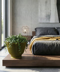 A green planter pot with a plant is placed on a wooden platform in a bedroom. The bed is in the background with gray bedding and a yellow blanket. A modern lamp sits on a nightstand next to the bed. A large window is on the left side of the image.