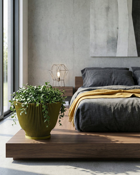 A green planter pot with a plant is placed on a wooden platform in a bedroom. The bed is in the background with gray bedding and a yellow blanket. A modern lamp sits on a nightstand next to the bed. A large window is on the left side of the image.