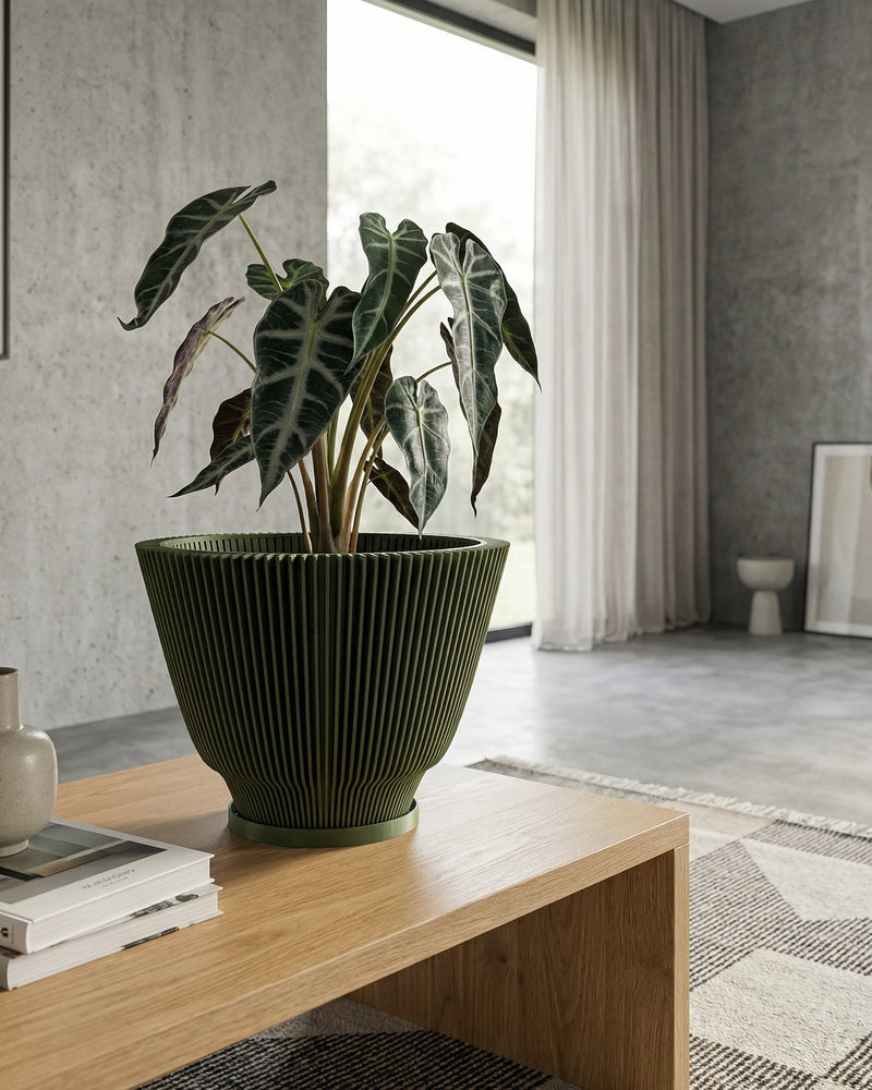 A green planter pot with a plant inside sits on a wooden table. The pot has a ribbed design. The table also has a vase and books on it. In the background, there is a window with a white curtain and a concrete wall.