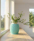 A light blue planter with a plant sits on a wooden windowsill in front of a window.