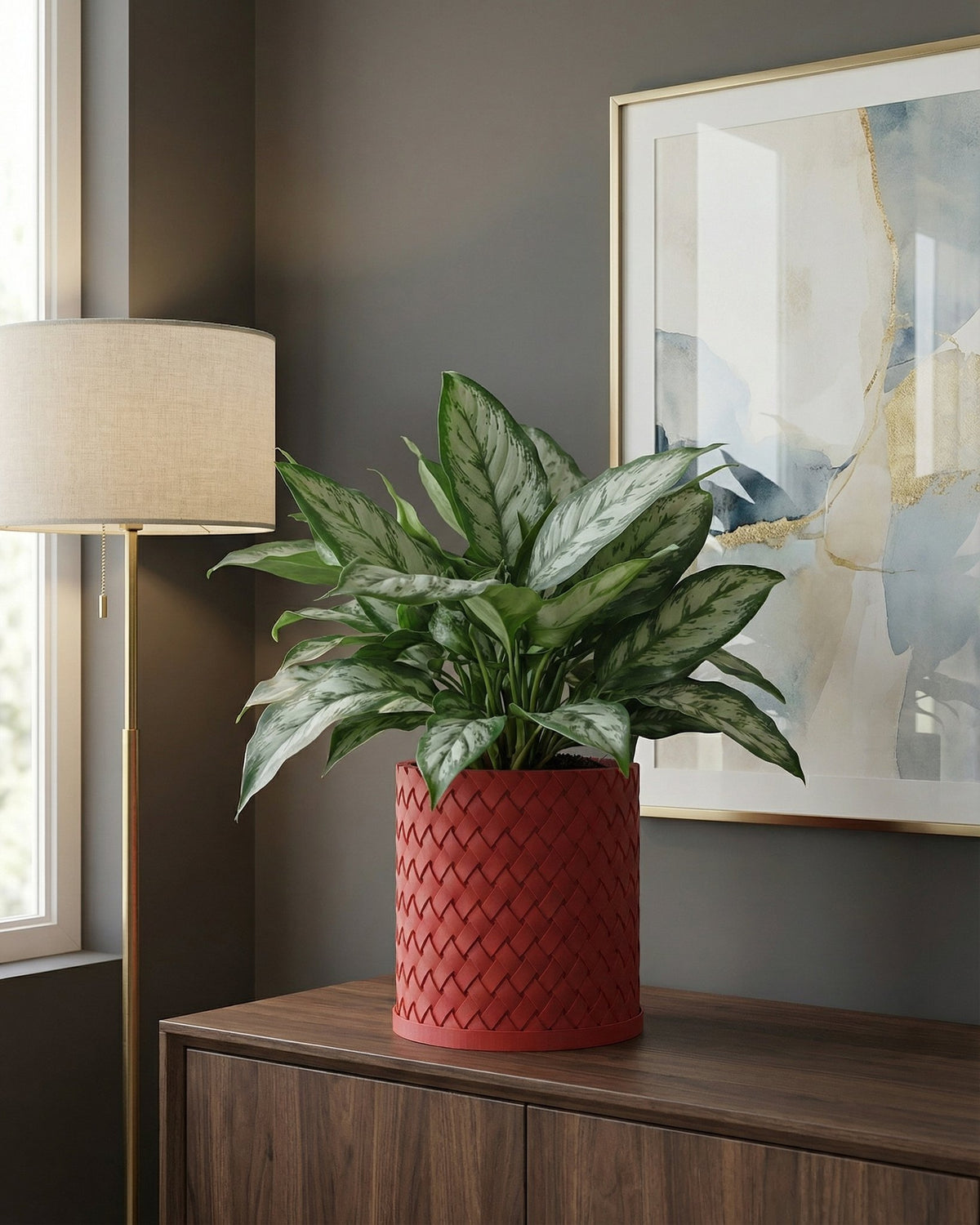 A red woven planter pot with a green plant sits on a wooden dresser, with a lamp and framed art in the background.