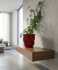 A red planter pot with a green plant sits on a wooden table next to a lamp. The pot has a textured design.
