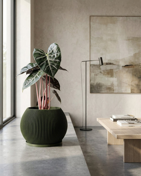 A green planter pot with a plant inside sits on a concrete surface near a window. A table and lamp are in the background.