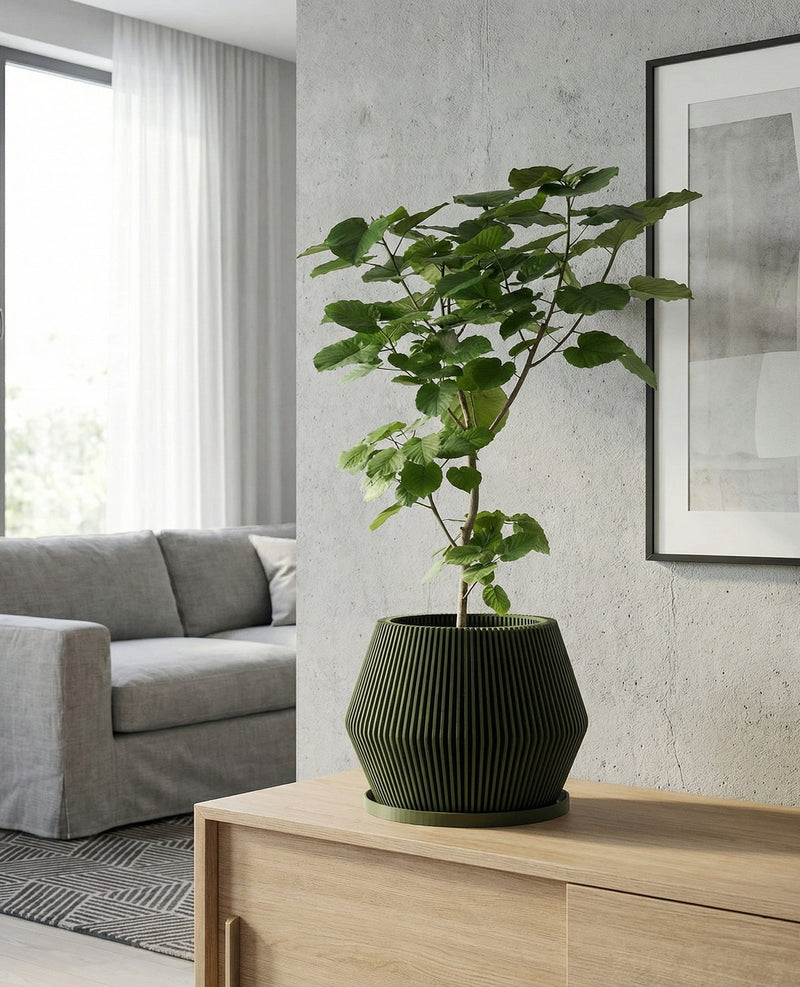 A green planter pot with a plant inside sits on a wooden table. The pot has a ribbed design. The table also has a vase and books on it. In the background, there is a window with a white curtain and a concrete wall.