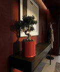 A red planter pot with a bonsai tree sits on a dark wood table against a red wall with a framed print.