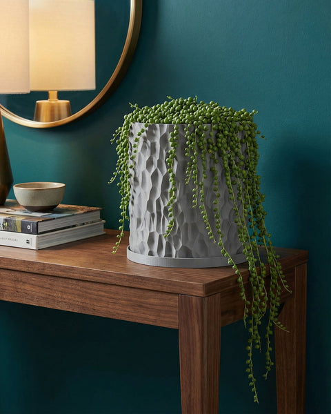 A gray planter pot with a string of pearls plant sits on a wooden table. The pot has a textured, geometric design.