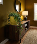 A yellow planter pot with a green plant on top of a wooden cabinet in a room with a mirror and a lamp.