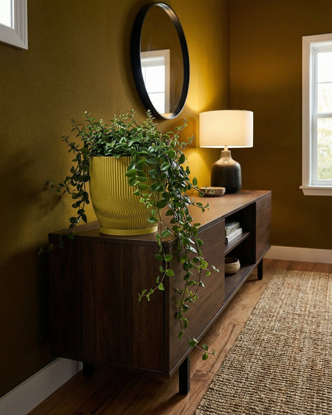 A yellow planter pot with a green plant on top of a wooden cabinet in a room with a mirror and a lamp.