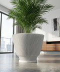 A large, white, fluted planter pot with a green plant inside sits on a gray concrete floor in a modern room. The pot has a unique, vertically ribbed design. A window with a city view is visible in the background.