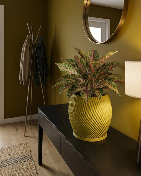 A yellow planter pot with a green and pink plant sits on a dark wood table in a room with olive green walls.