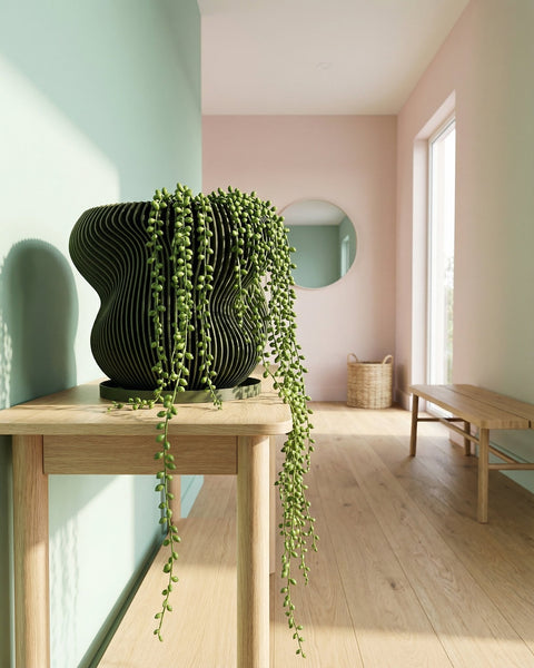 A green planter pot with a plant inside sits on a concrete surface near a window. A table and lamp are in the background.