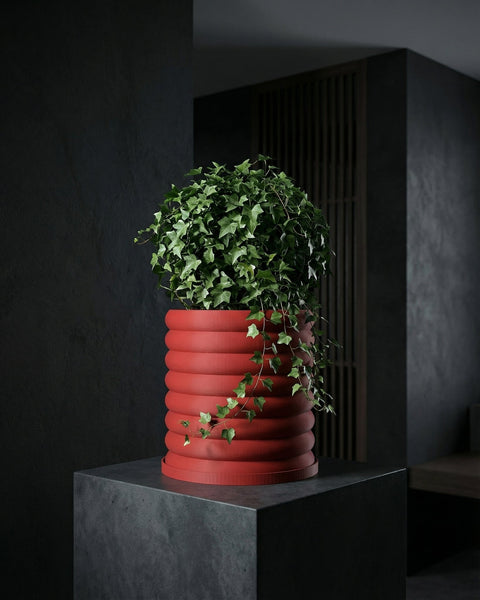 A red, ribbed planter pot sits atop a dark gray pedestal, filled with lush green ivy. The background is a dark, textured wall.