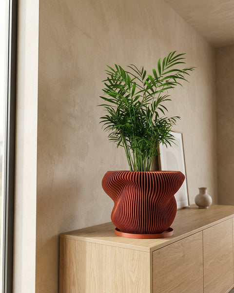 A plant in a ribbed, round, terracotta-colored planter sits on a wooden windowsill next to a bright window.