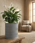 A peace lily in a gray planter pot with a wavy design sits on a wooden table in a cozy room.