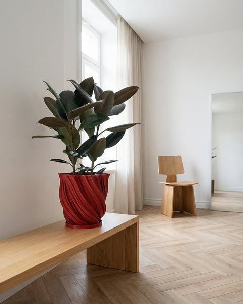 A red planter pot with a green plant sits on a wooden shelf in front of a window. The pot has a spiral design.