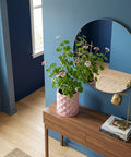 A pink planter pot with a plant on a wooden table, next to a round mirror and a lamp. The pot has a geometric pattern.