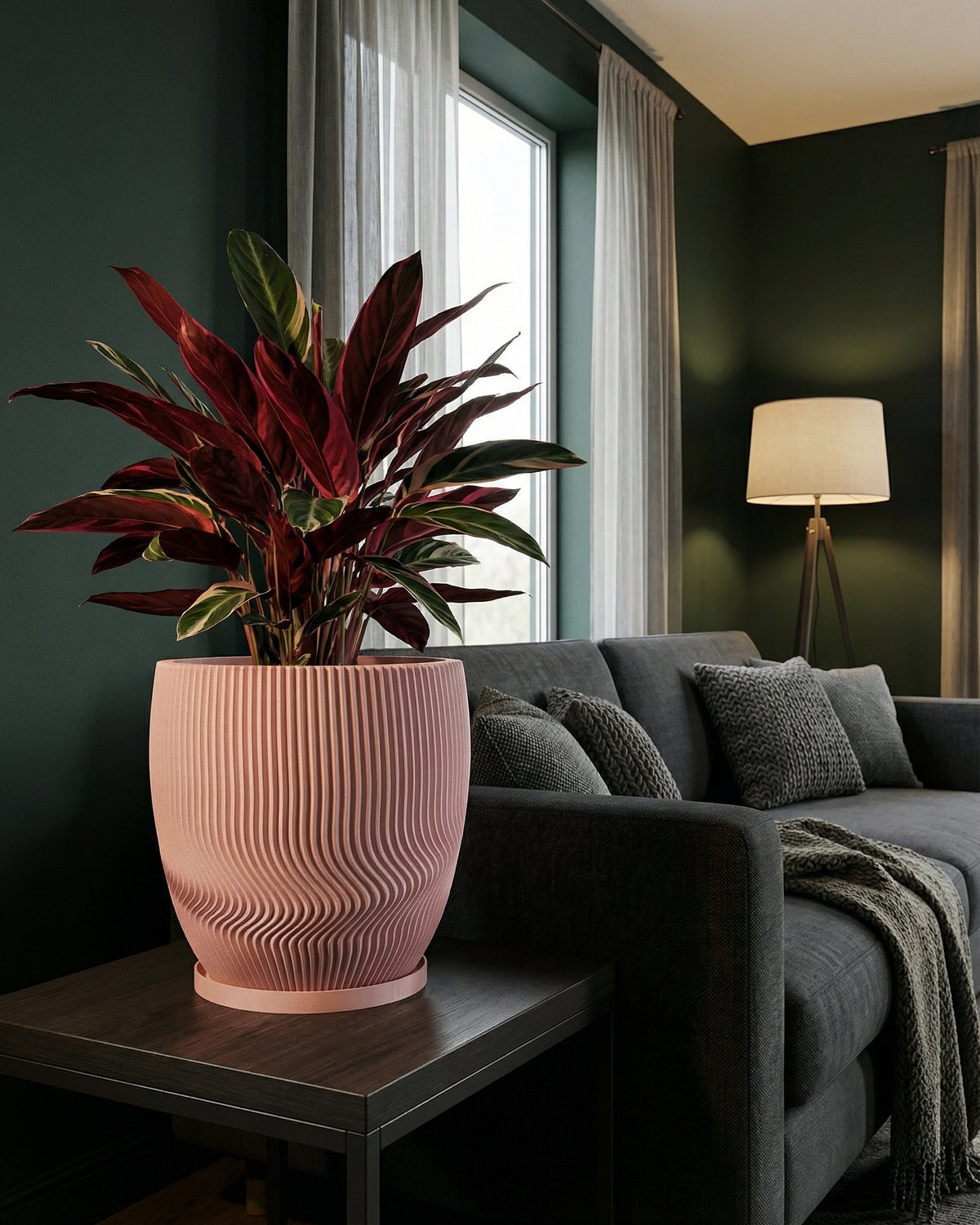 A pink planter pot with a ribbed design sits on a dark wood table next to a gray couch in a living room. A plant with red and green leaves is in the pot.