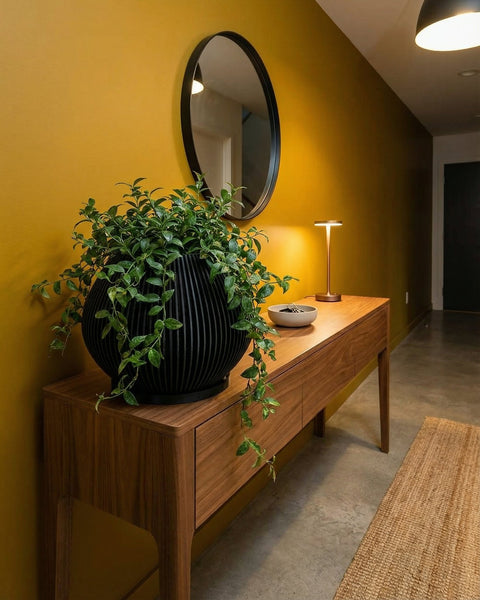 A Monstera plant in a black, vertically ribbed planter pot sits on a gray floor in a modern room. A large window is to the left, and a light wood paneled wall is behind the plant. A white sofa and abstract art are in the background.