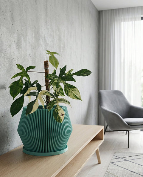 A green planter pot with a plant inside sits on top of a wooden cabinet. The cabinet is in a room with a concrete wall and a painting on the wall. A floor lamp is next to the cabinet.