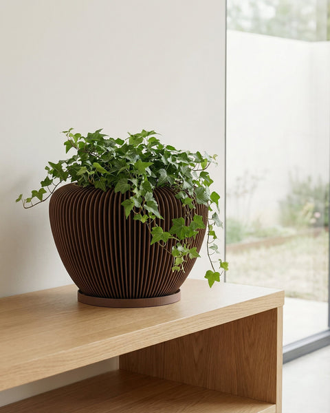 A brown, decorative planter pot with a green ivy plant sits on a light wood shelf against a white wall.