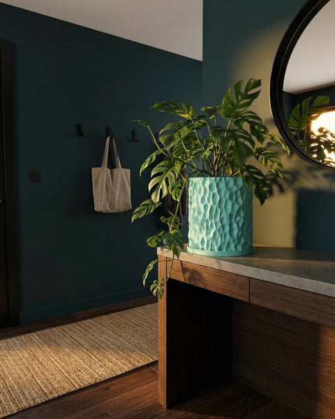 A monstera plant in a textured, light blue planter pot sits on a wooden table in a room with dark teal walls.