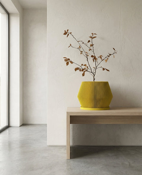 A yellow planter pot with a plant inside sits on a gray concrete cube. The pot has a fluted design and the plant has green and silver leaves. The cube is in a room with a concrete floor and a window.
