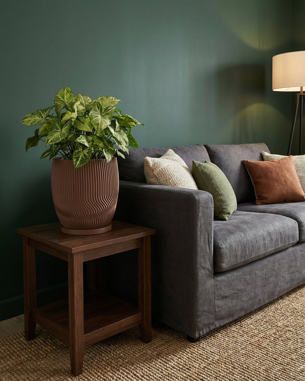A brown planter pot with a plant sits on a wooden side table next to a gray couch with pillows.