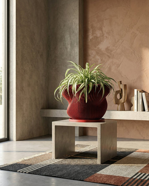 A red, round, ribbed planter pot with a green plant inside is placed on a wooden floor in a room with a window.