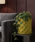 A yellow planter pot with a plant in it sits on a dark brown table next to a gray couch. The background is a dark brown wall.