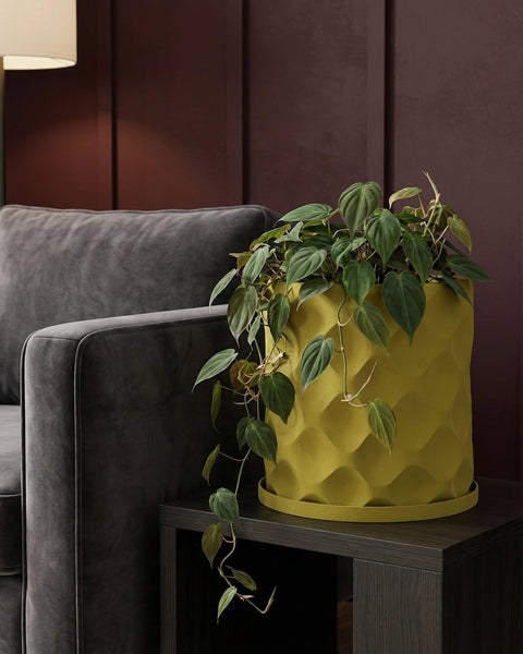 A yellow planter pot with a plant in it sits on a dark brown table next to a gray couch. The background is a dark brown wall.