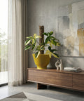A yellow planter pot with a plant inside sits on a wooden cabinet in a modern living room. The pot is ribbed and has a saucer. The plant has green and yellow leaves and is supported by a moss pole. A white sculpture and two books are on the cabinet next to the pot. A painting hangs on the wall behind the cabinet. A sheer curtain covers the window to the left.