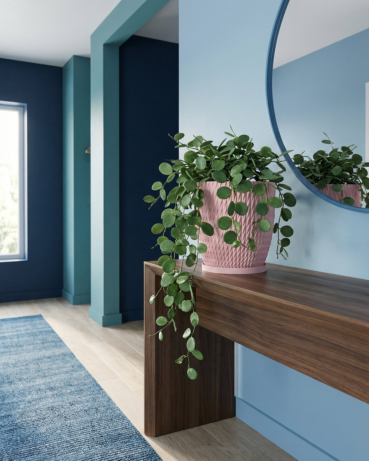 A pink planter pot with a green plant sits on a wooden table in a blue room with a round mirror.