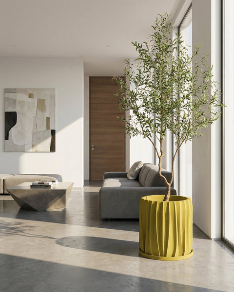 A yellow textured planter pot with a plant next to a gray couch and a round rug in a room with dark green walls.