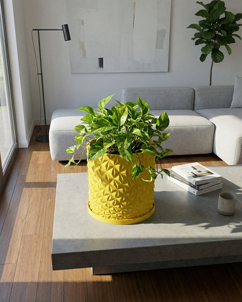 A vibrant yellow planter pot with a geometric design sits on a gray coffee table, holding a lush green plant. Books and a mug are nearby.