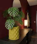A monstera plant in a yellow geometric planter sits on a dark wood cabinet in a room with red walls. A mirror and lamp are nearby.
