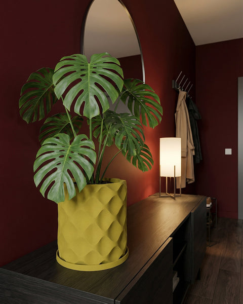 A monstera plant in a yellow geometric planter sits on a dark wood cabinet in a room with red walls. A mirror and lamp are nearby.