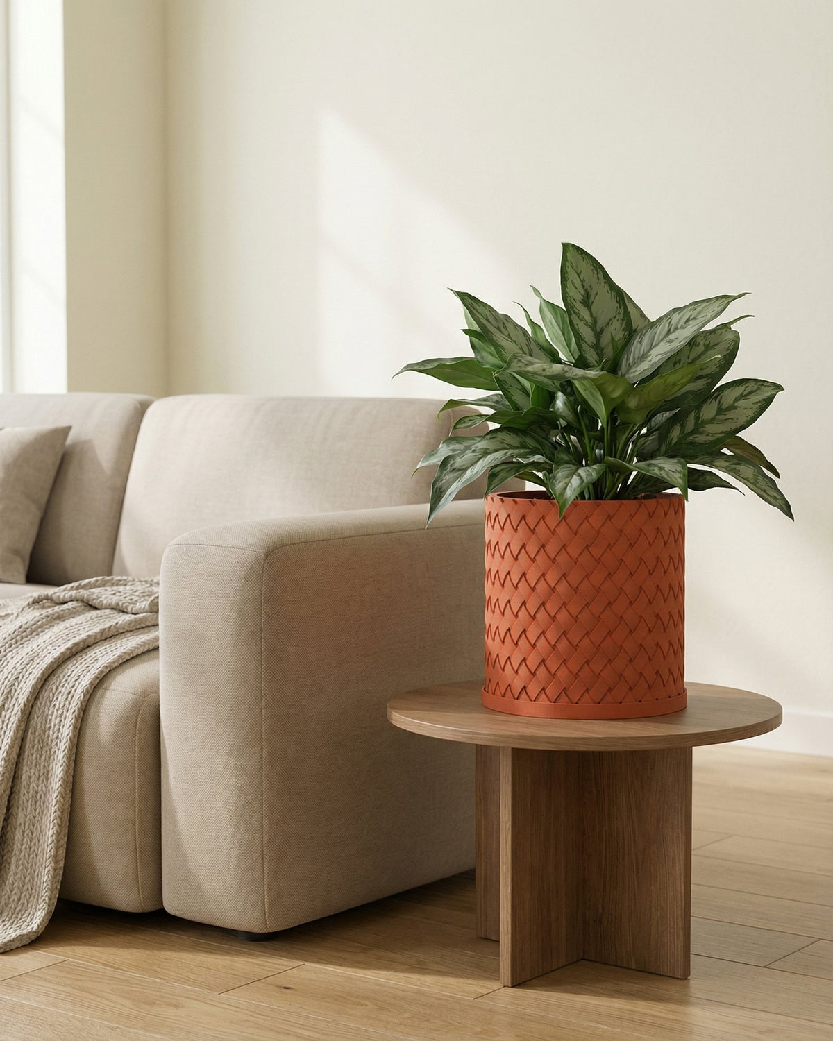 A woven, terracotta-colored planter pot sits on a wooden side table next to a beige couch with a throw blanket.