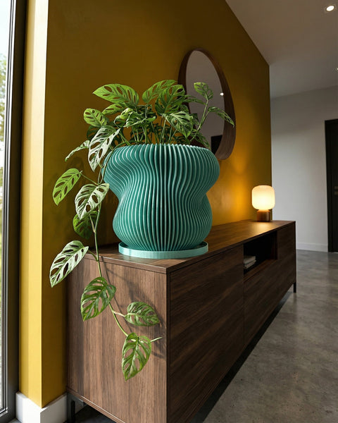 A green ribbed planter pot sits on a wooden cabinet, holding a plant with pink and dark green leaves, next to books and a geometric object.