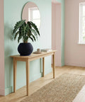 A dark blue ribbed planter pot sits on a light wood cabinet, holding a vibrant green and pink houseplant. The background features a concrete wall and a bright, airy room with sheer curtains.