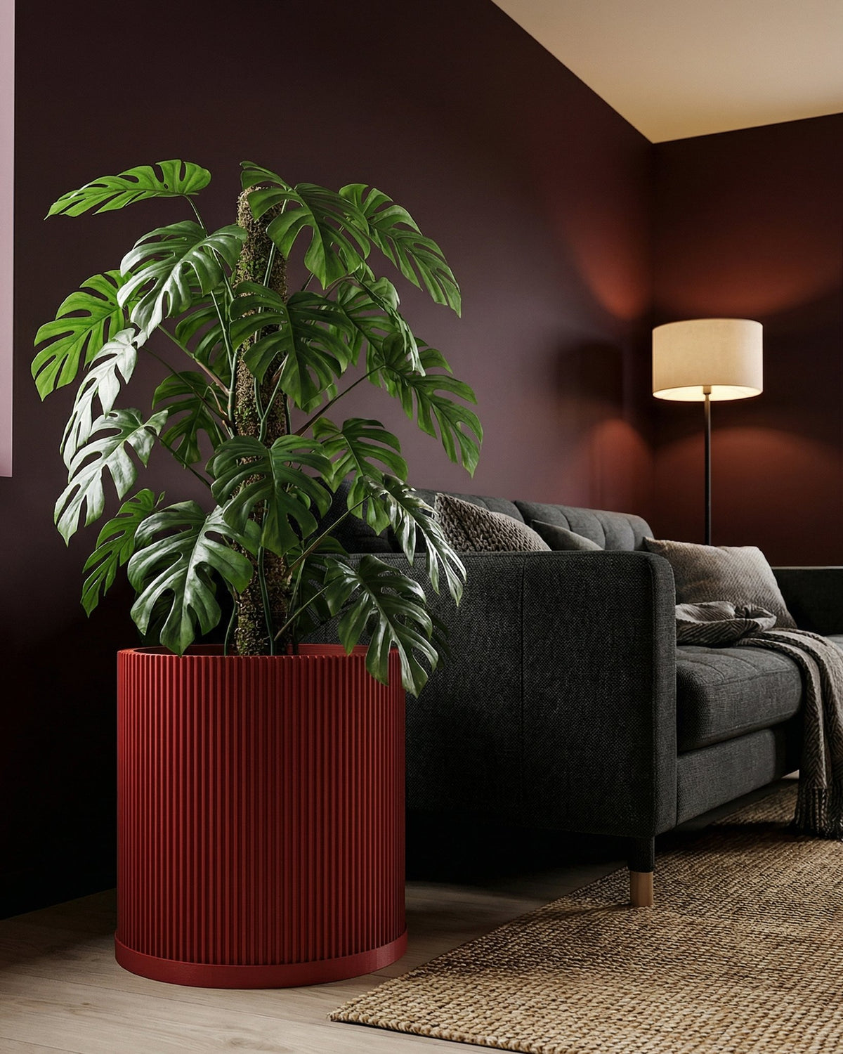 A red planter pot with a monstera plant sits next to a gray couch in a room with dark red walls and a floor lamp.
