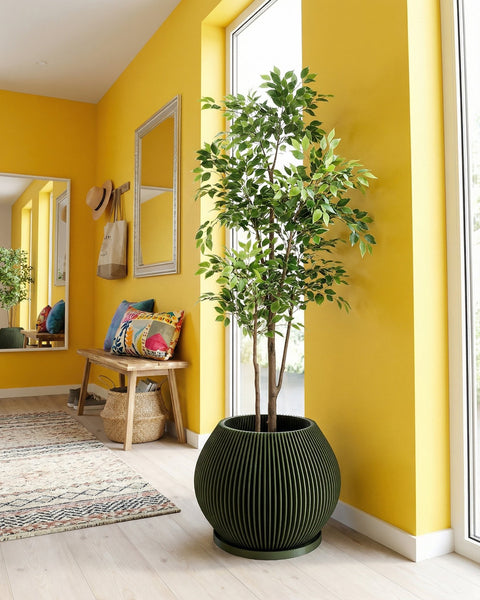 A green planter pot with a plant inside sits on a wooden table. The pot has a ribbed design. The table also has a vase and books on it. In the background, there is a window with a white curtain and a concrete wall.
