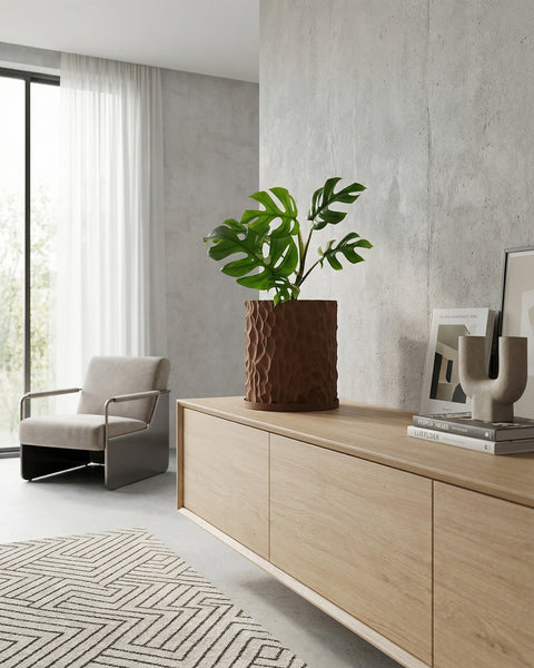 A brown textured planter pot with a green plant sits on a light wood cabinet in a modern living room.